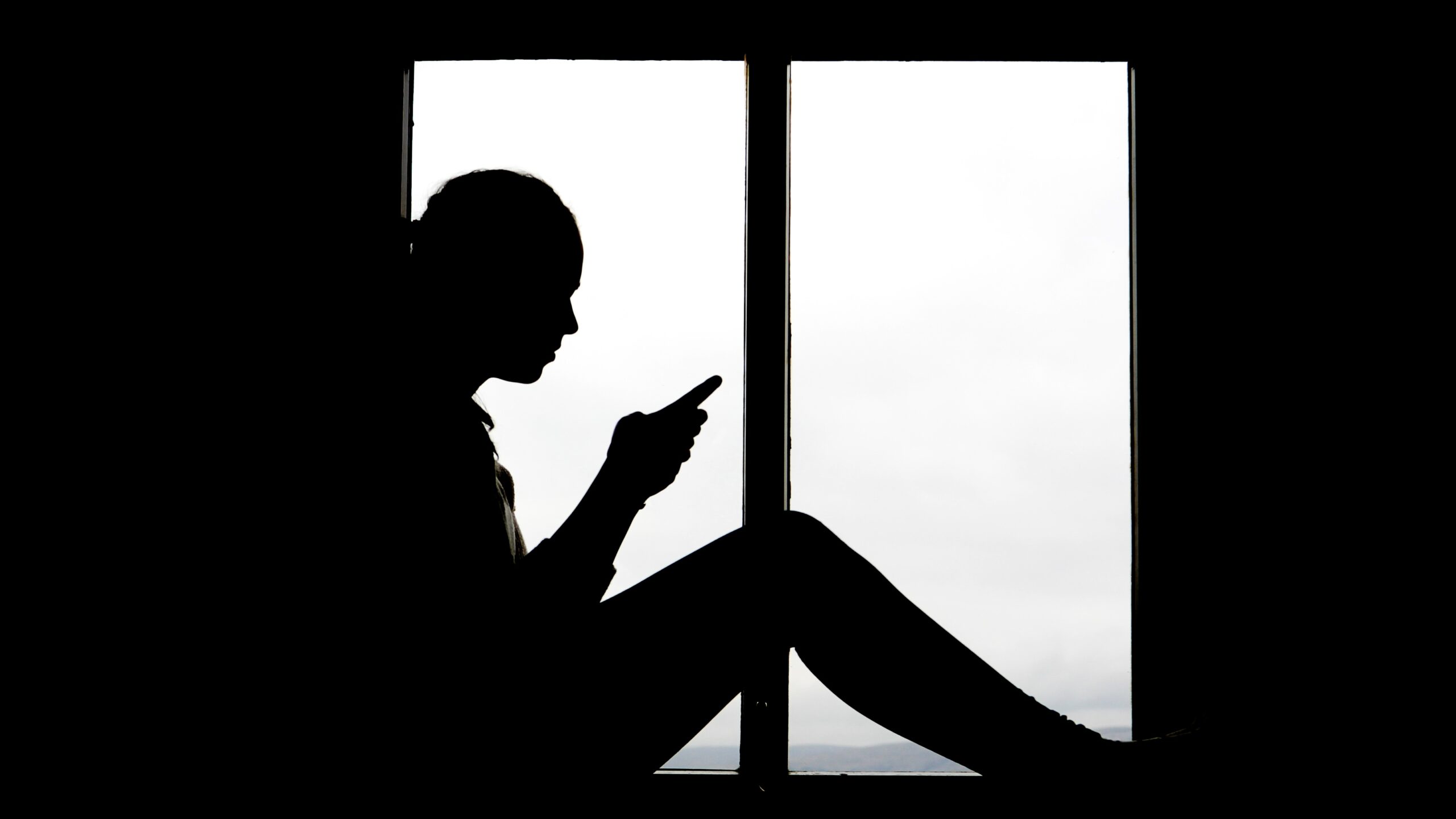 Silhouette of 16 year old girl texting backlit at window.